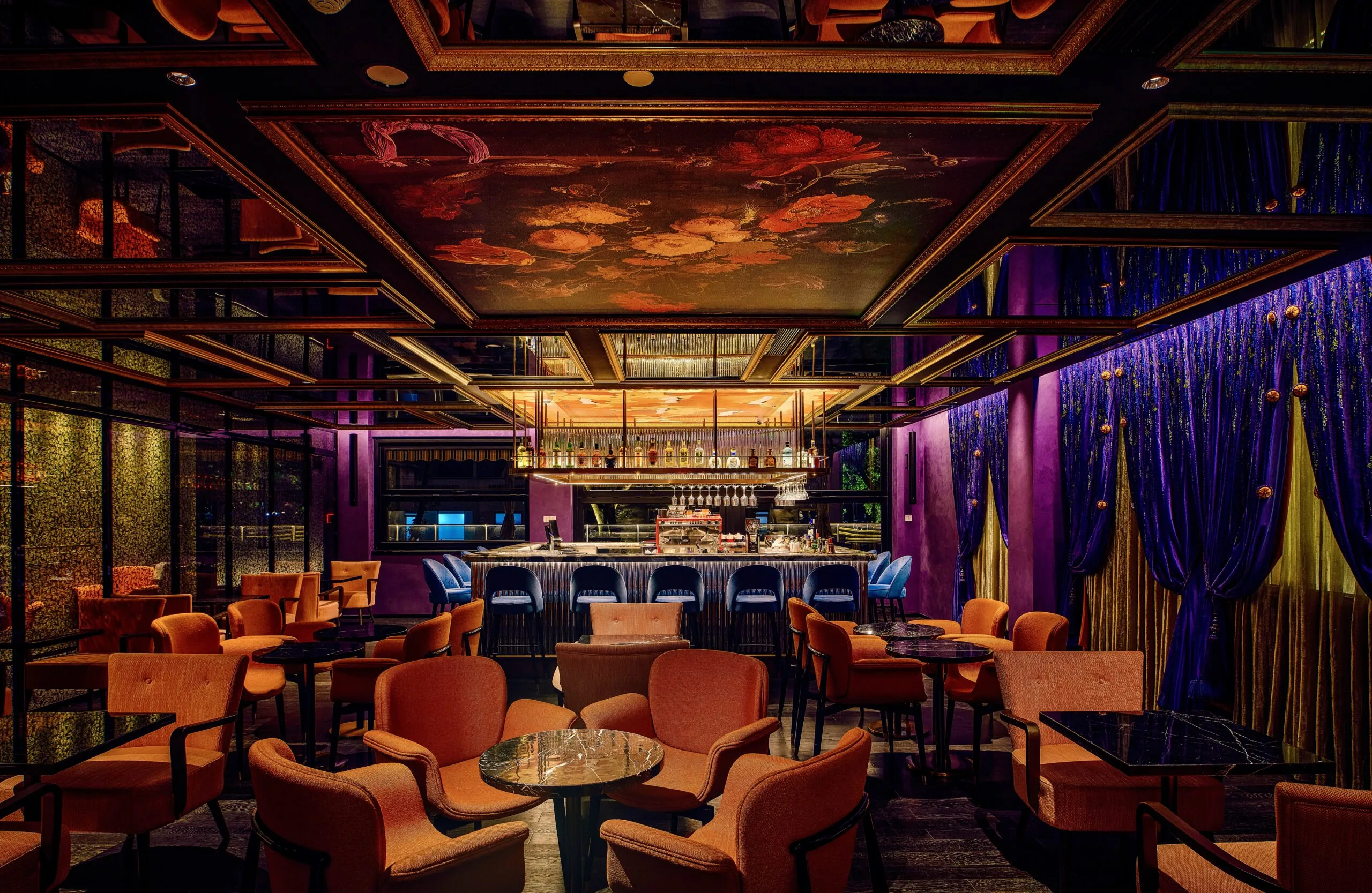 Luxurious bar with plush orange seating, a mirrored ceiling with floral art, and opulent purple velvet curtains.