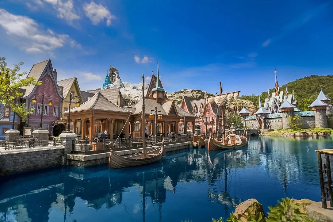 Two wooden boats dock at a waterfront village with a castle in the background under a blue sky.