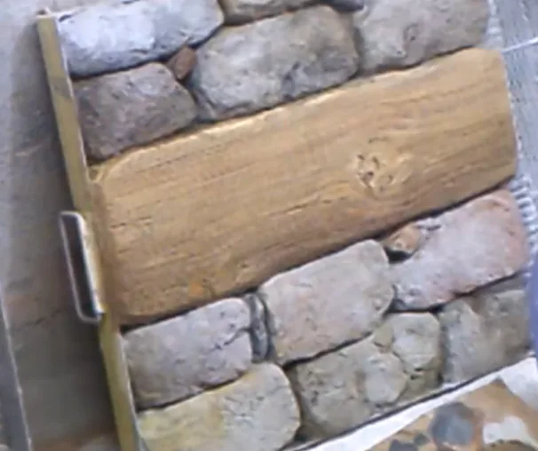 A textured wooden beam is set against a rustic stone wall with a metallic bracket.
